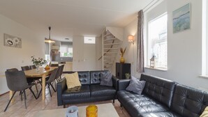 A modern living room with a black leather sofa, a dining table with chairs, and a kitchen in the background. - Holiday Home in Zeeland With Garden (Wemeldinge)