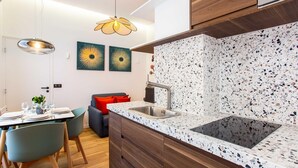 A modern kitchen with a granite countertop, stainless steel sink, and wooden cabinets. There are two wall-mounted pendant lights and a dining area with a table and chairs.