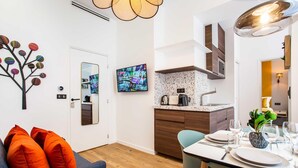 A modern living room with a sofa, orange cushions, a dining table set for two, a kitchen with a granite countertop, and a wall-mounted TV.