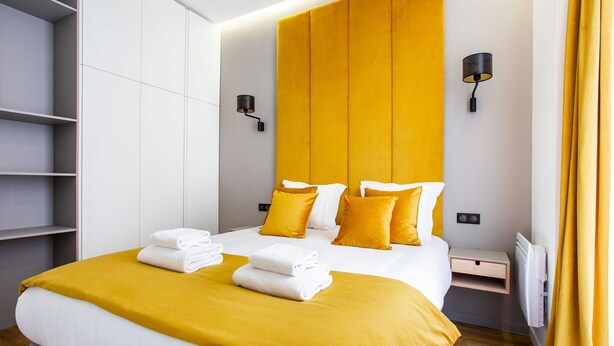 A modern hotel room with a large bed, a bedside table, a built-in shelf, and a wall-mounted lamp.