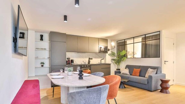 Een moderne woonkamer met een grijze bank, een ronde eettafel met stoelen, een rode bank en een groot raam met gordijnen.