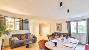 Een moderne woonkamer met een grijze bank, een ronde eettafel gedekt voor vier personen en een groot raam met gordijnen.