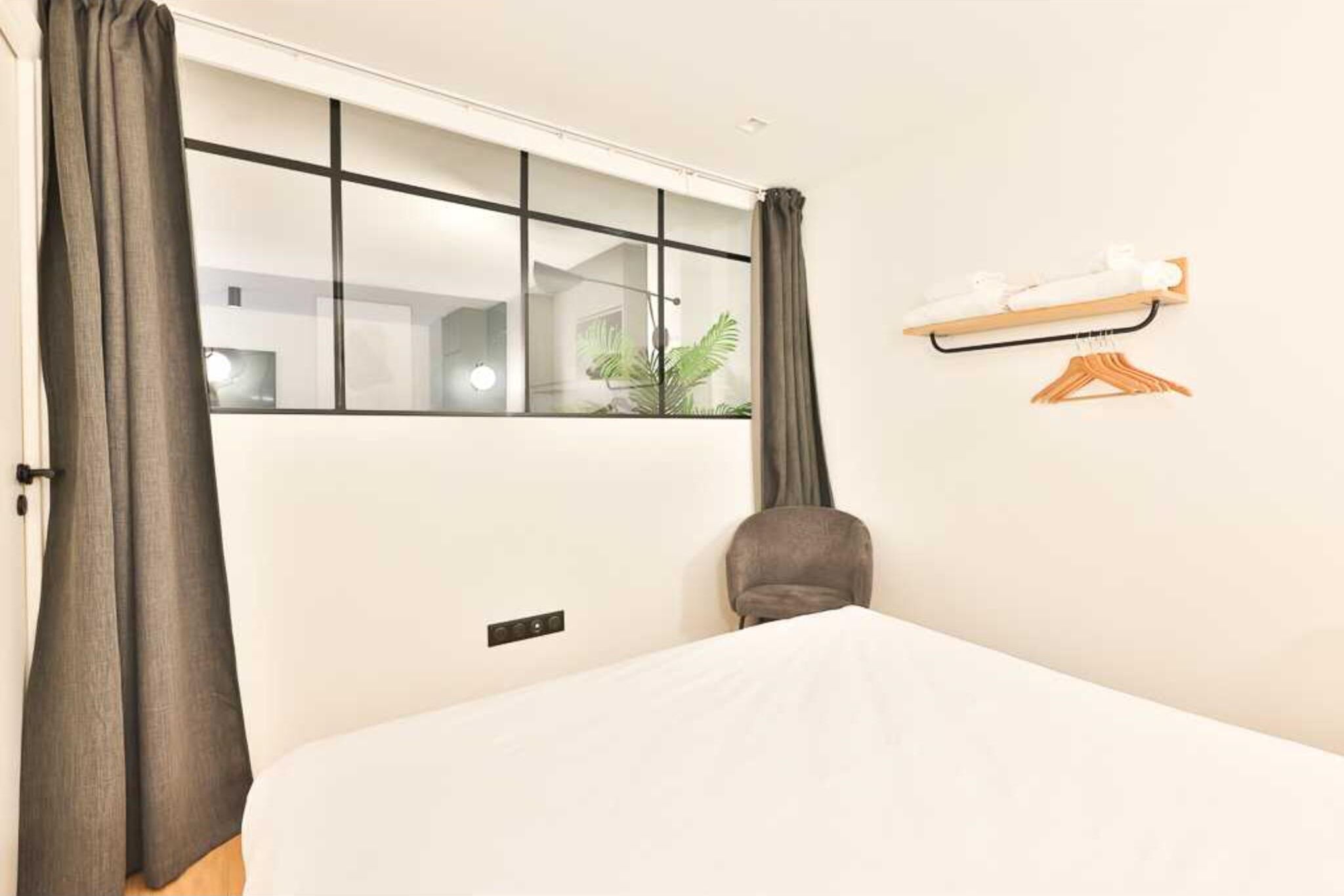 Quarto de hotel com uma janela grande, cama, cadeira, prateleira pequena com toalhas e uma planta.