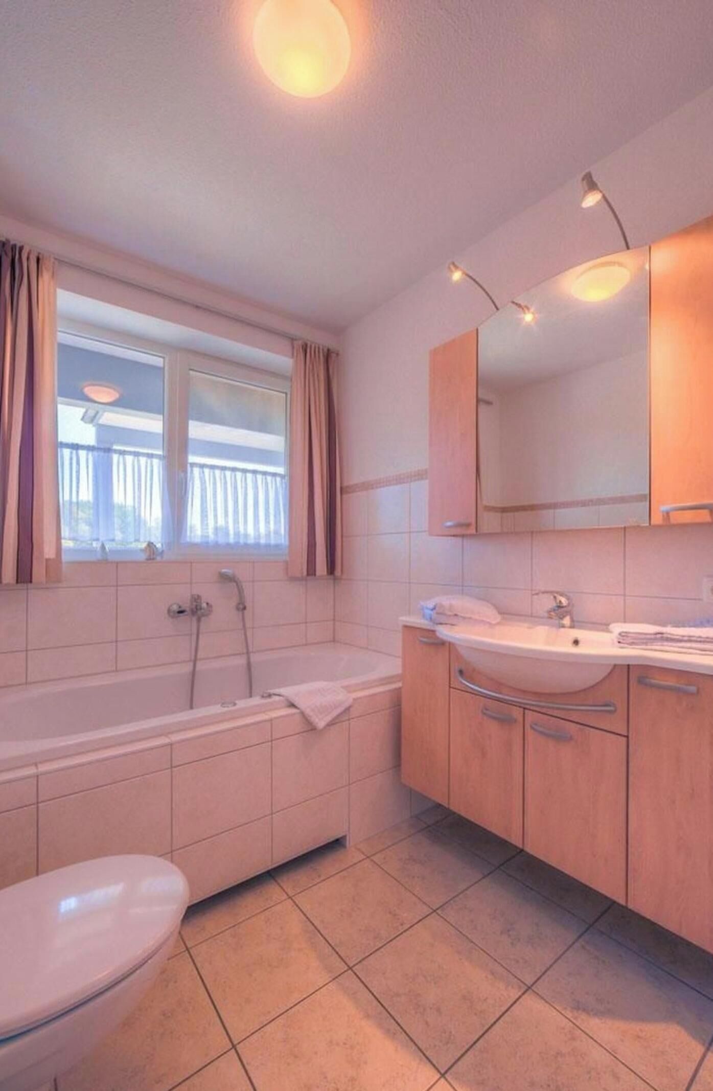 Un baño con bañera, lavamanos e inodoro. Cuenta con una ventana con cortinas y suelo de baldosas.