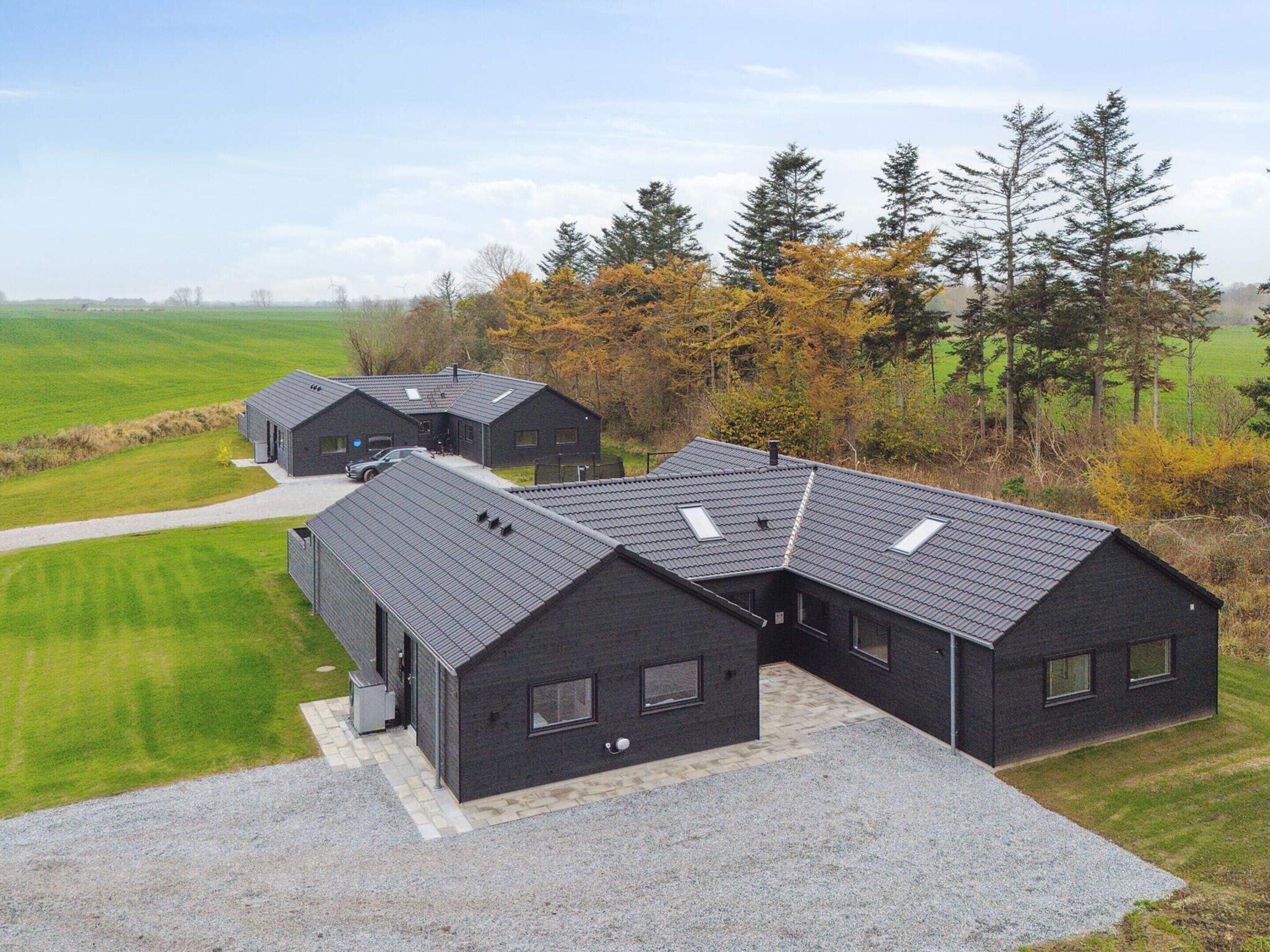A modern, dark-colored building complex with multiple sections, surrounded by a rural landscape with fields and trees.