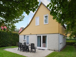 A two-story house with a yellow exterior and white brick lower level. There is a patio with a table and chairs, and a sliding glass door leading to a covered outdoor area.