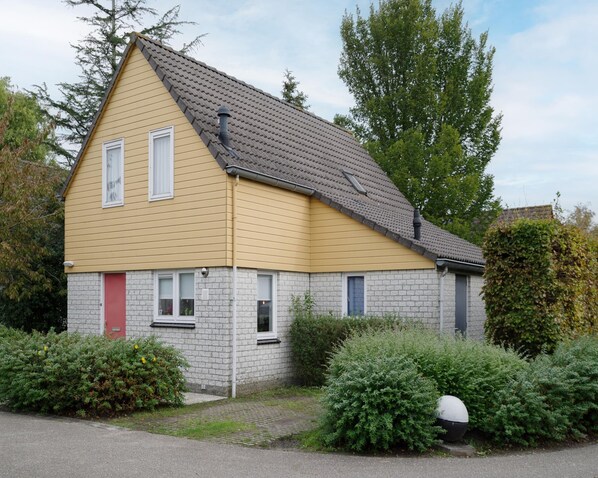 A two-story house with a yellow exterior and grey brick foundation. It features a red door, two windows, and a chimney.