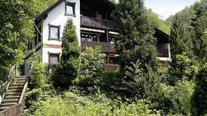 A two-story chalet with a balcony, surrounded by dense forest.