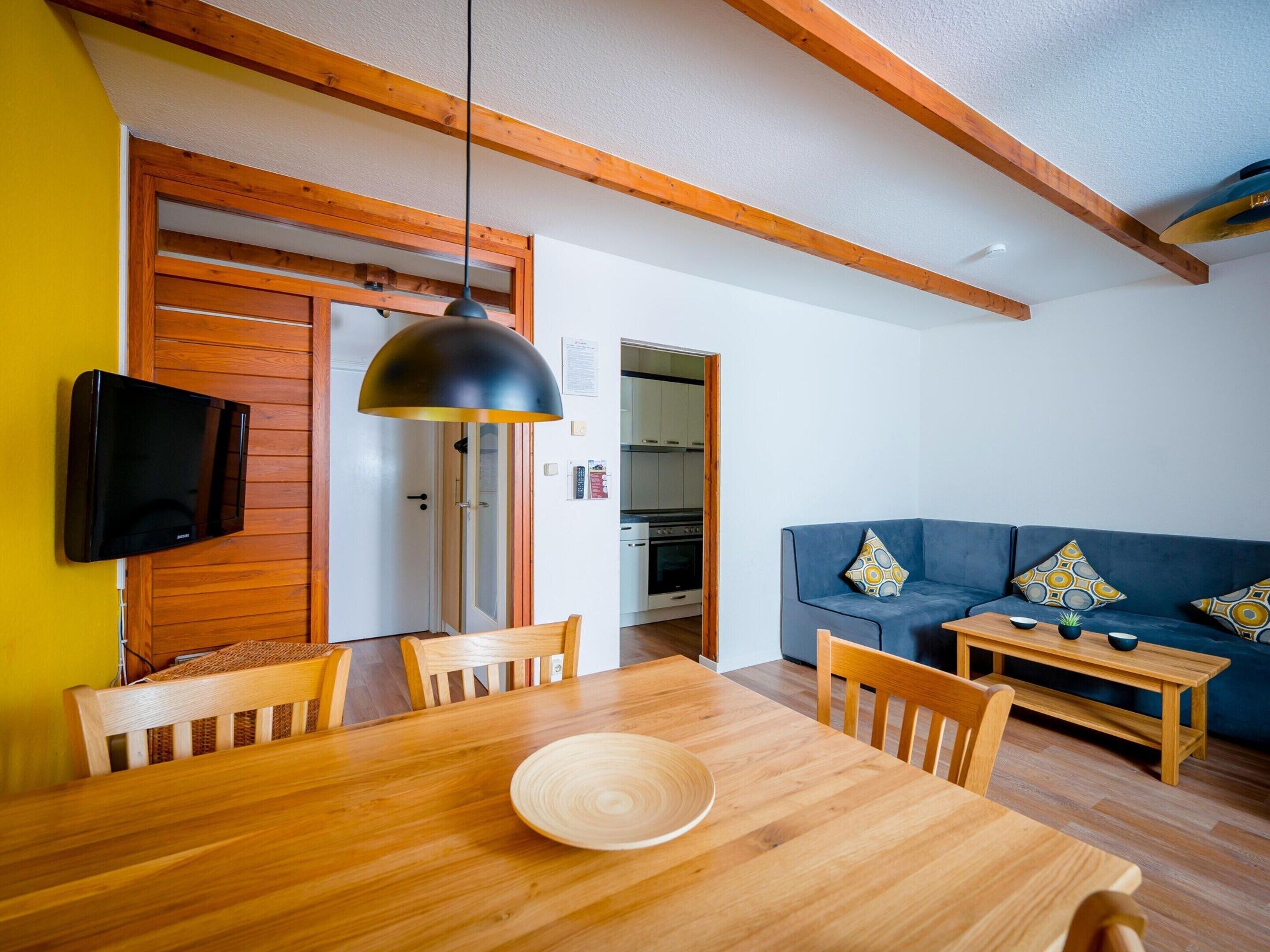 Zona pranzo con tavolo e sedie in legno, lampada a sospensione nera e televisore fissato a una parete di legno.