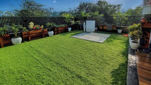 A well-maintained backyard with artificial grass, wooden planters with various plants, a white outdoor seating area, and a black metal fence. - Fun O Peace (Noida)