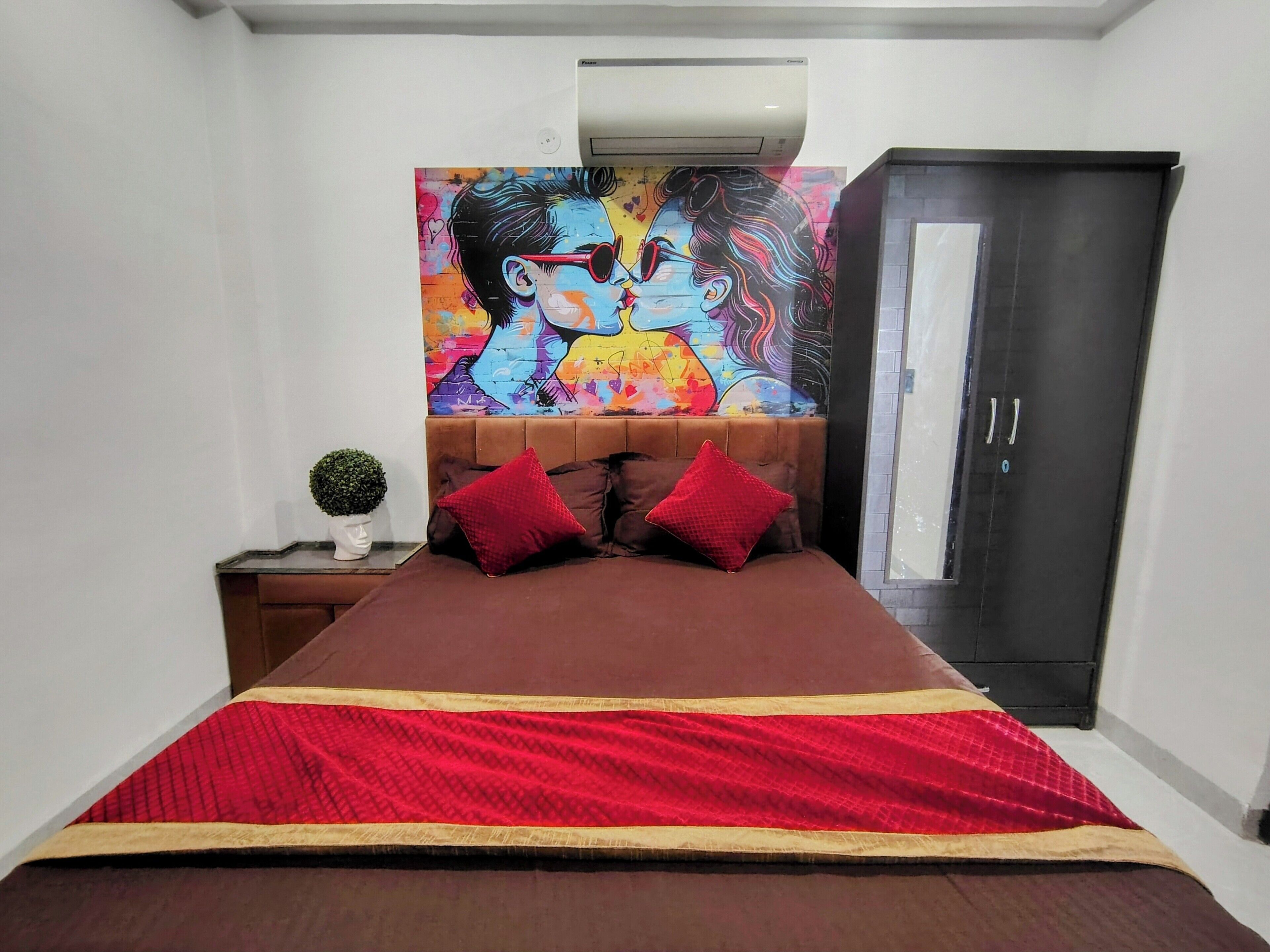 A bedroom with a bed, a nightstand with a plant, a colorful painting above the bed, and a wardrobe.