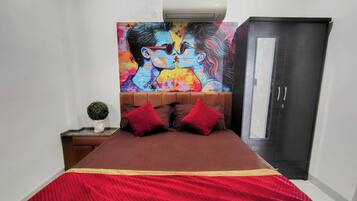 A bedroom with a bed, a nightstand with a plant, a colorful painting above the bed, and a wardrobe.