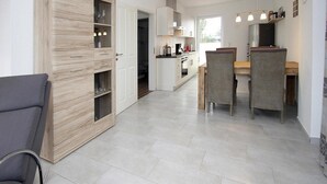 A modern kitchen with white cabinets, a dining area with wicker chairs, and a grey sofa. - Ferienhaus Albatros in Nessmersiel (Dornum)