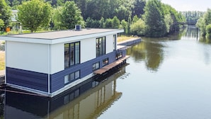 A modern houseboat with a flat roof, large windows, and a dock extending into a calm river. - Houseboat With Private Roofed Terrace (Zeewolde)