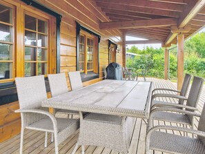 A covered outdoor dining area with a long table and wicker chairs, surrounded by wooden walls and large windows.