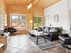 A spacious living room with a wooden ceiling, large windows, a black leather sofa, a coffee table, and a desk with a chair.
