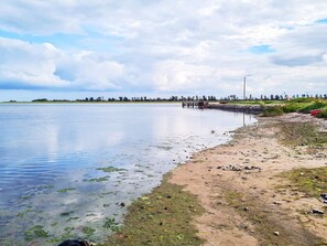 A calm lake with a sandy shore, a wooden dock, and a distant tree line.