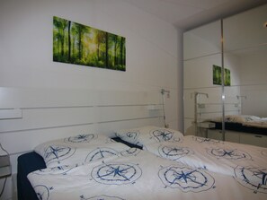 A bedroom with a bed, a bedside table, a mirror, and a painting of a forest. - Ferienwohnung auf Fehmarn in Orther Reede (Fehmarn)