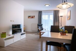 A modern living room with a flat-screen TV, a dining table with chairs, a lamp, and a window with curtains. - Ferienwohnung auf Fehmarn in Orther Reede (Fehmarn)