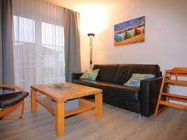 Ein Wohnzimmer mit einer Couch, einem Holztisch und einem Sessel. An der Wand hängt ein Gemälde und es steht eine Lampe bereit.