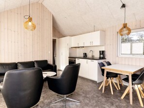 A modern kitchen with white cabinets, a black countertop, and a coffee maker. There is a dining area with a wooden table and black chairs. - 5 Star Holiday Home in Lokken (Lokken)