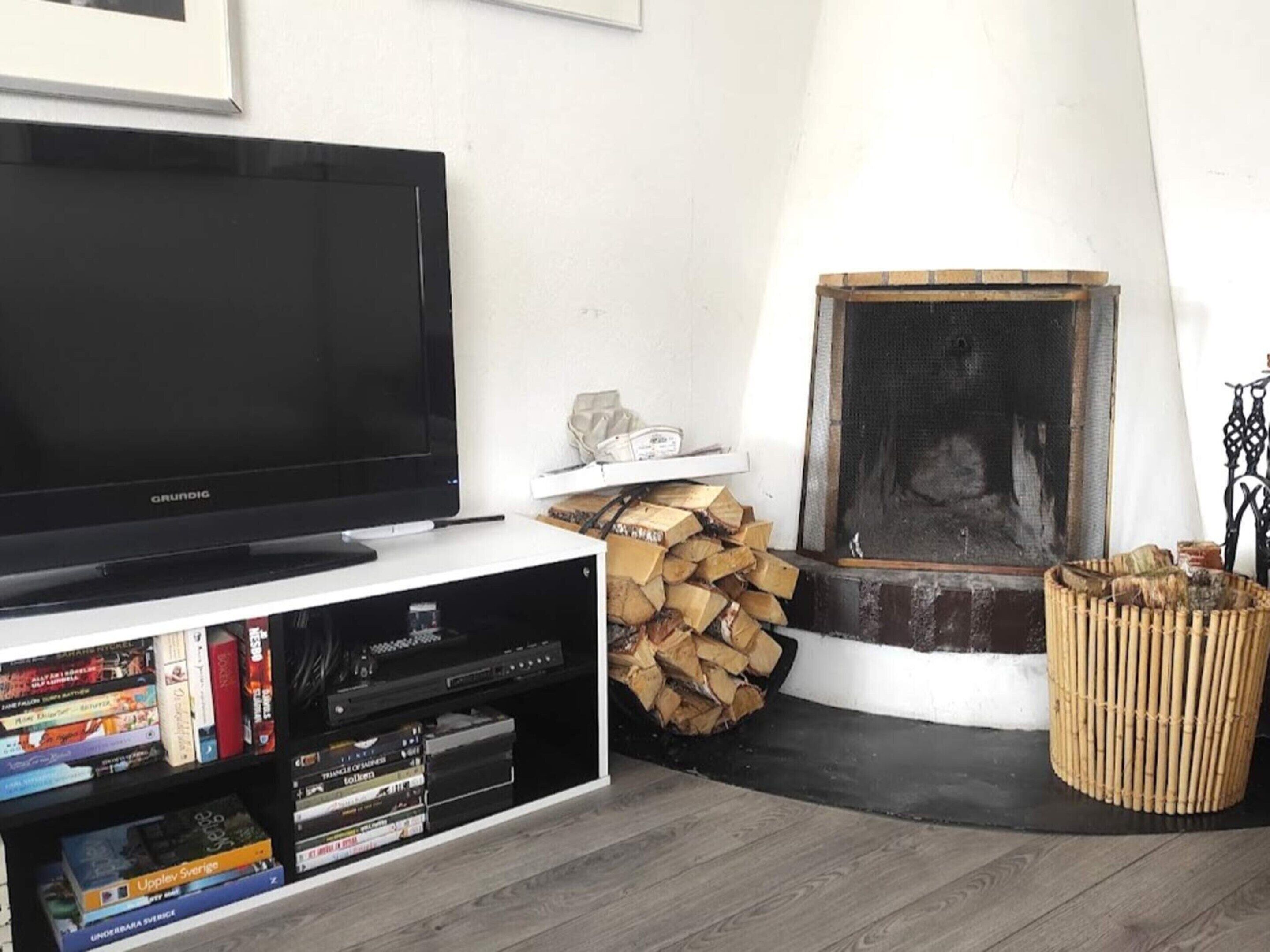Una sala de estar con televisor sobre un mueble bajo, chimenea, leña apilada y una estantería con varios objetos.