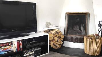 Una sala de estar con televisor sobre un mueble bajo, chimenea, leña apilada y una estantería con varios objetos.