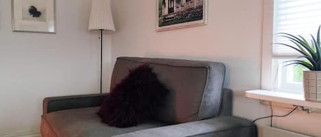 Een woonkamer met een grijze bank, een zwart kussen, een ingelijst schilderij, een vloerlamp en een raam met een plant.