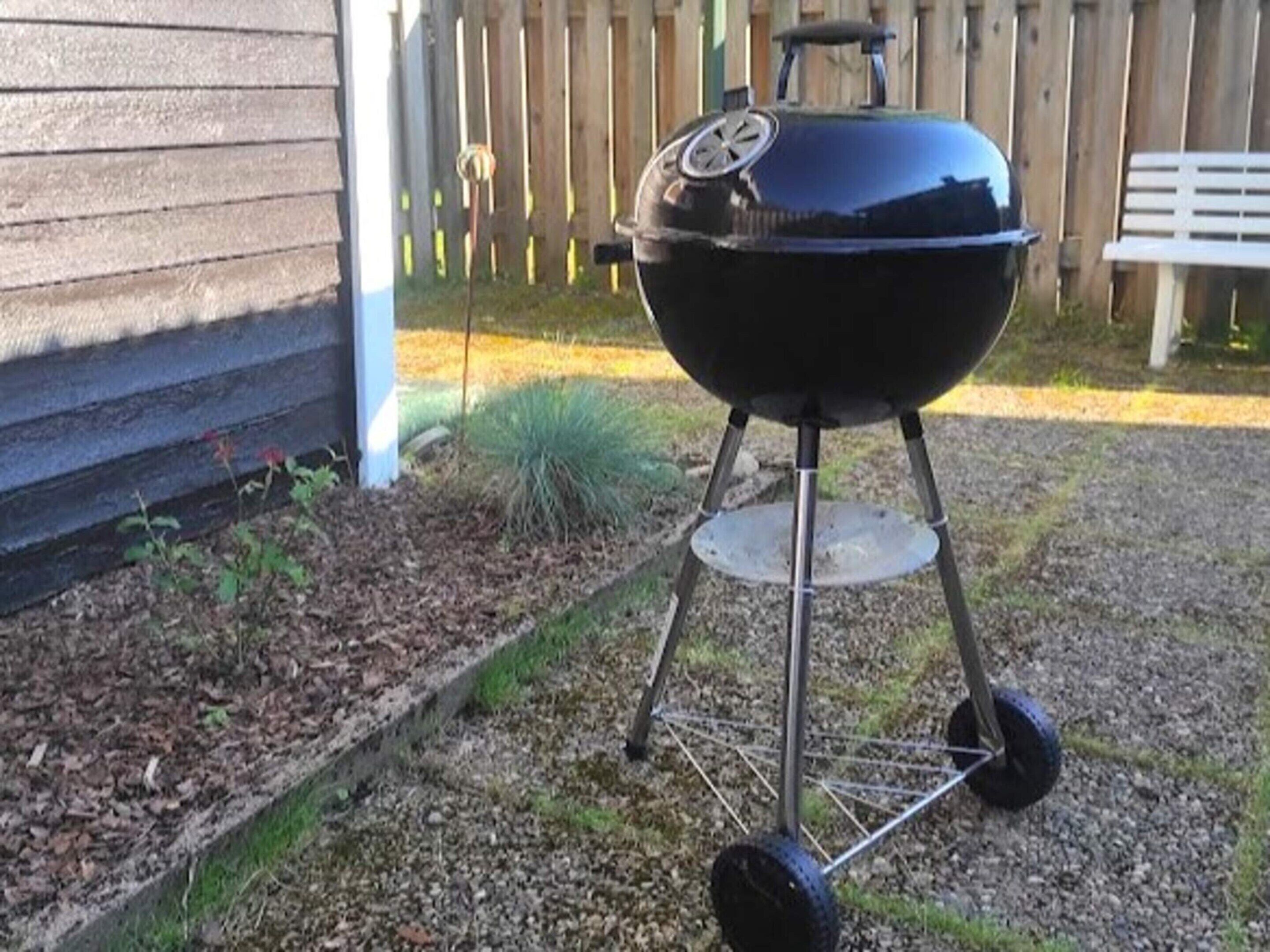 Una parrilla para barbacoa con ruedas, ubicada sobre grava cerca de una valla de madera y una casa.