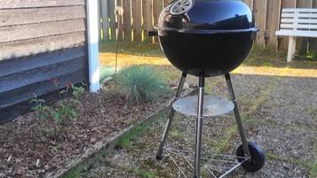 Una parrilla para barbacoa con ruedas, ubicada sobre grava cerca de una valla de madera y una casa.