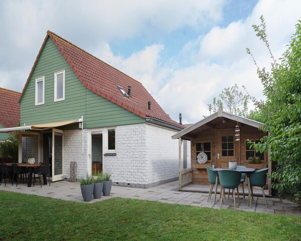 Une maison à deux étages avec un toit végétalisé et des murs blancs, dotée d’un patio avec des meubles d’extérieur et des plantes en pot.
