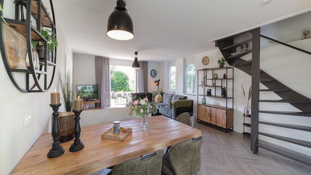 A modern living space with a wooden dining table, green upholstered chairs, a black pendant light, a staircase, and a bookshelf. - Holiday Home in Quiet Park With Garden (Wemeldinge)