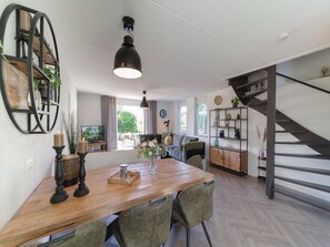 A modern living space with a wooden dining table, green upholstered chairs, a black pendant light, a staircase, and a bookshelf. - Holiday Home in Quiet Park With Garden (Wemeldinge)