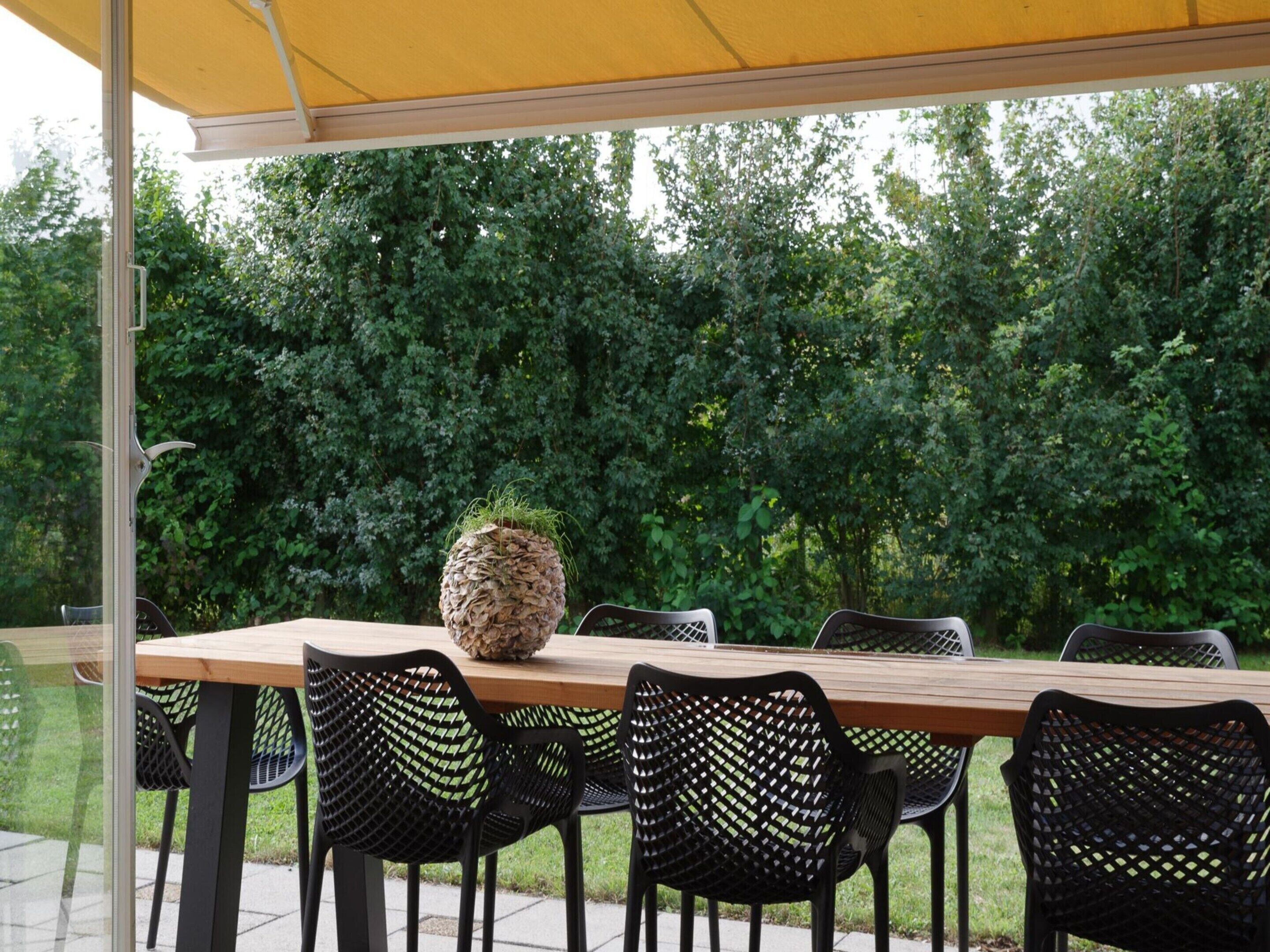 An outdoor patio with a wooden table and black mesh chairs, surrounded by lush greenery.
