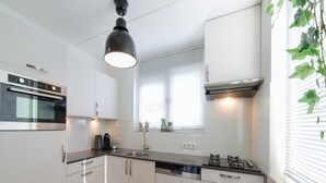A modern kitchen with white cabinets, stainless steel appliances, a black pendant light, and a window with blinds. - Holiday Home in Quiet Park With Garden (Wemeldinge)