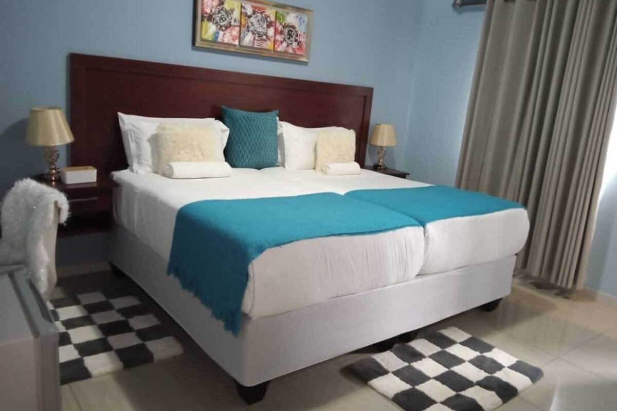 A neatly made bed with a blue blanket, white pillows, and a wooden headboard. There are two bedside lamps and a checkered rug on the floor.