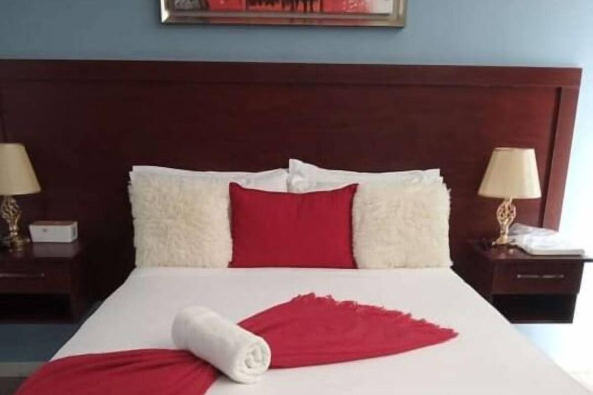 A neatly made bed with white linens, a red throw blanket, and a rolled white towel.