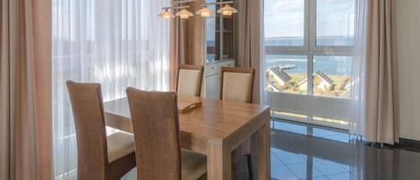 A modern dining area with a wooden table and wicker chairs, large windows with curtains, and a view of the sea.