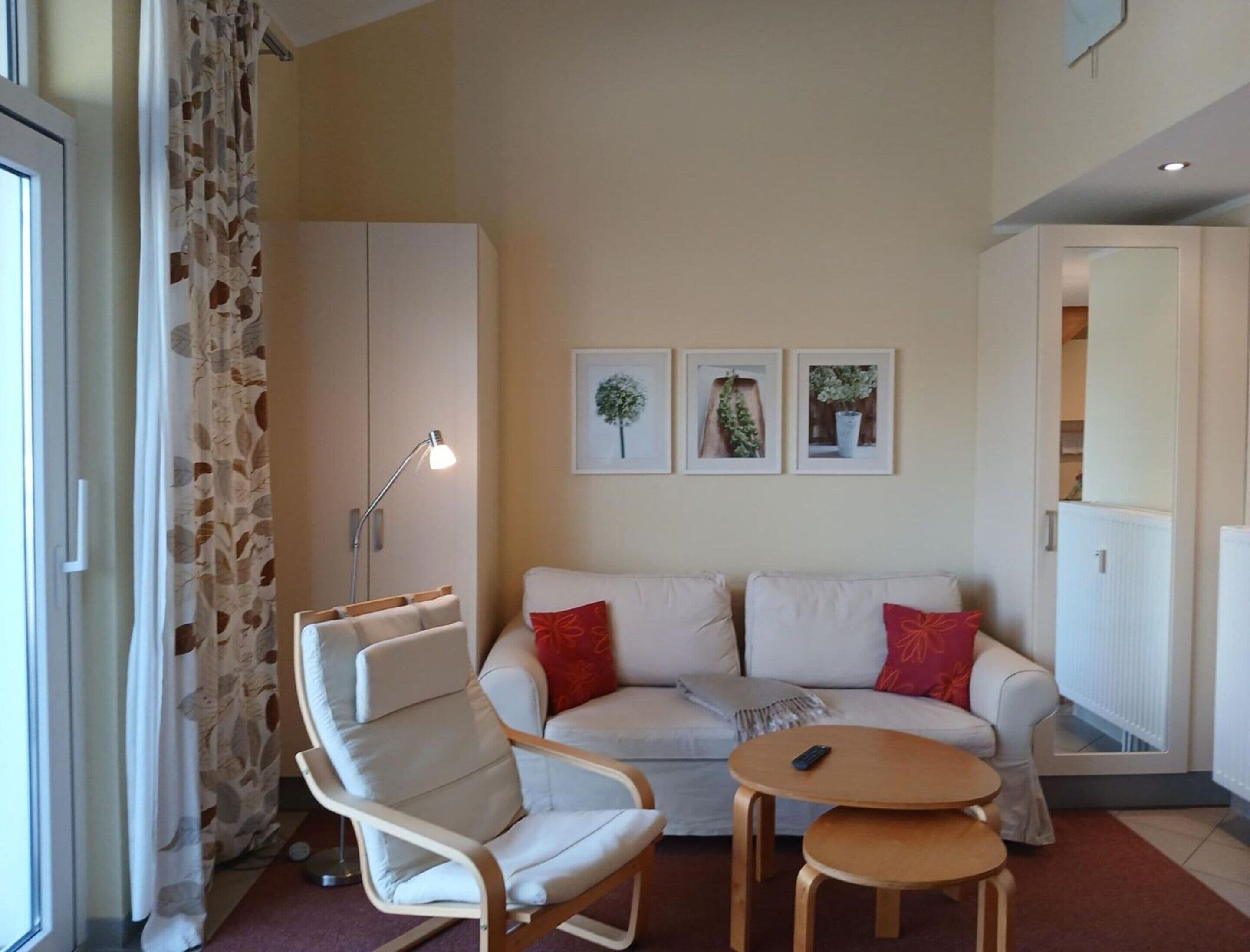 Ein Wohnzimmer mit einer Couch, einem Sessel, einem kleinen Tisch und einer Lampe. An der Wand hängen drei gerahmte Bilder.
