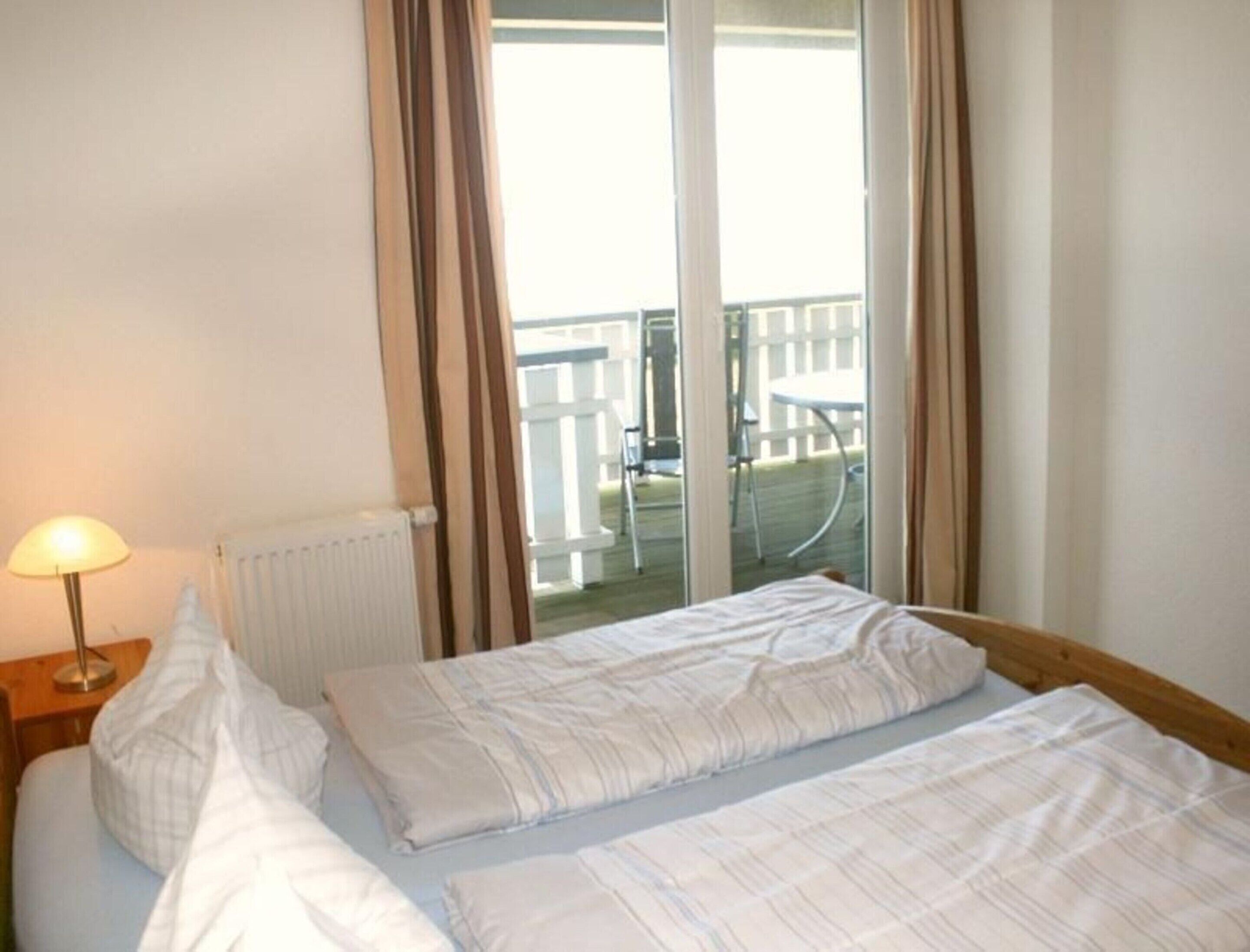 A hotel room with two single beds, a radiator, a lamp, and a view of a balcony with outdoor furniture.