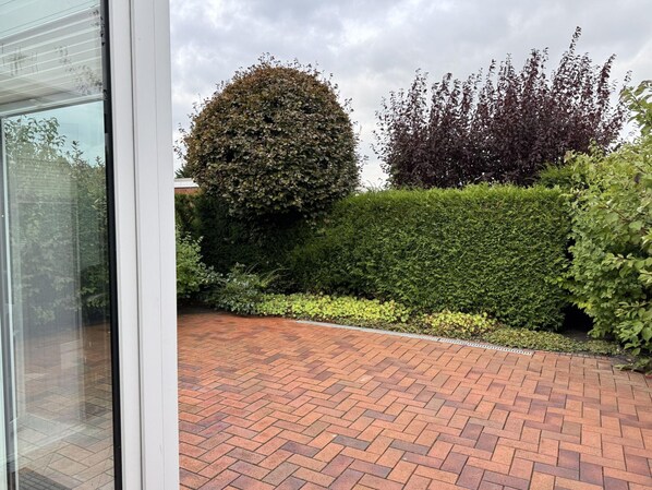 A paved courtyard with a brick pattern, surrounded by well-maintained hedges and a tall, dense tree. - Bungalow, Dornumersiel (Dornum)