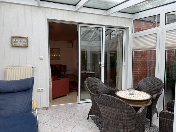 A sunlit patio with a sliding glass door leading to an indoor dining area, wicker furniture, and a tiled floor. - Bungalow, Dornumersiel (Dornum)