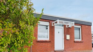 A red brick building with a white door and windows, a small porch, and a tree in the foreground. - Bungalow, Dornumersiel (Dornum)