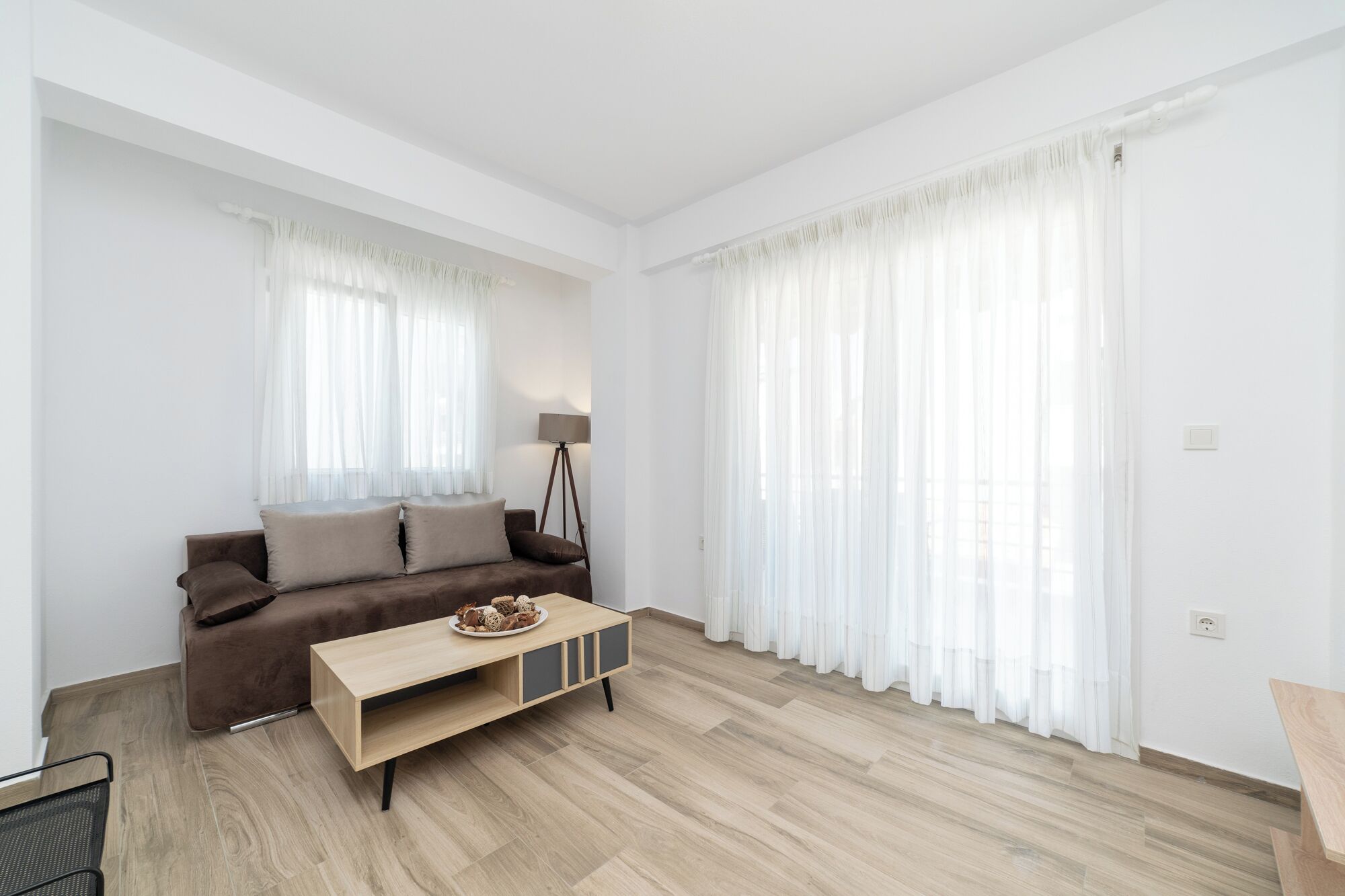 Een moderne woonkamer met een bruine bank, een houten salontafel en lichte, witte gordijnen.