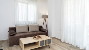 A modern living room with a brown sofa, a wooden coffee table with decorative items, and sheer white curtains. - Amarylis Residential Complex-One Bedroom Apartment (Kavala)