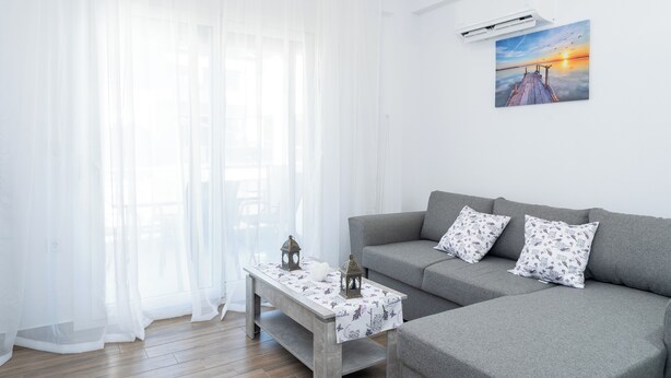 A modern living room with a grey sofa, two patterned pillows, a wooden coffee table with a white tablecloth, and a painting of a sunset on the wall. - Amarylis Residential Complex-One Bedroom Apartment (Kavala)