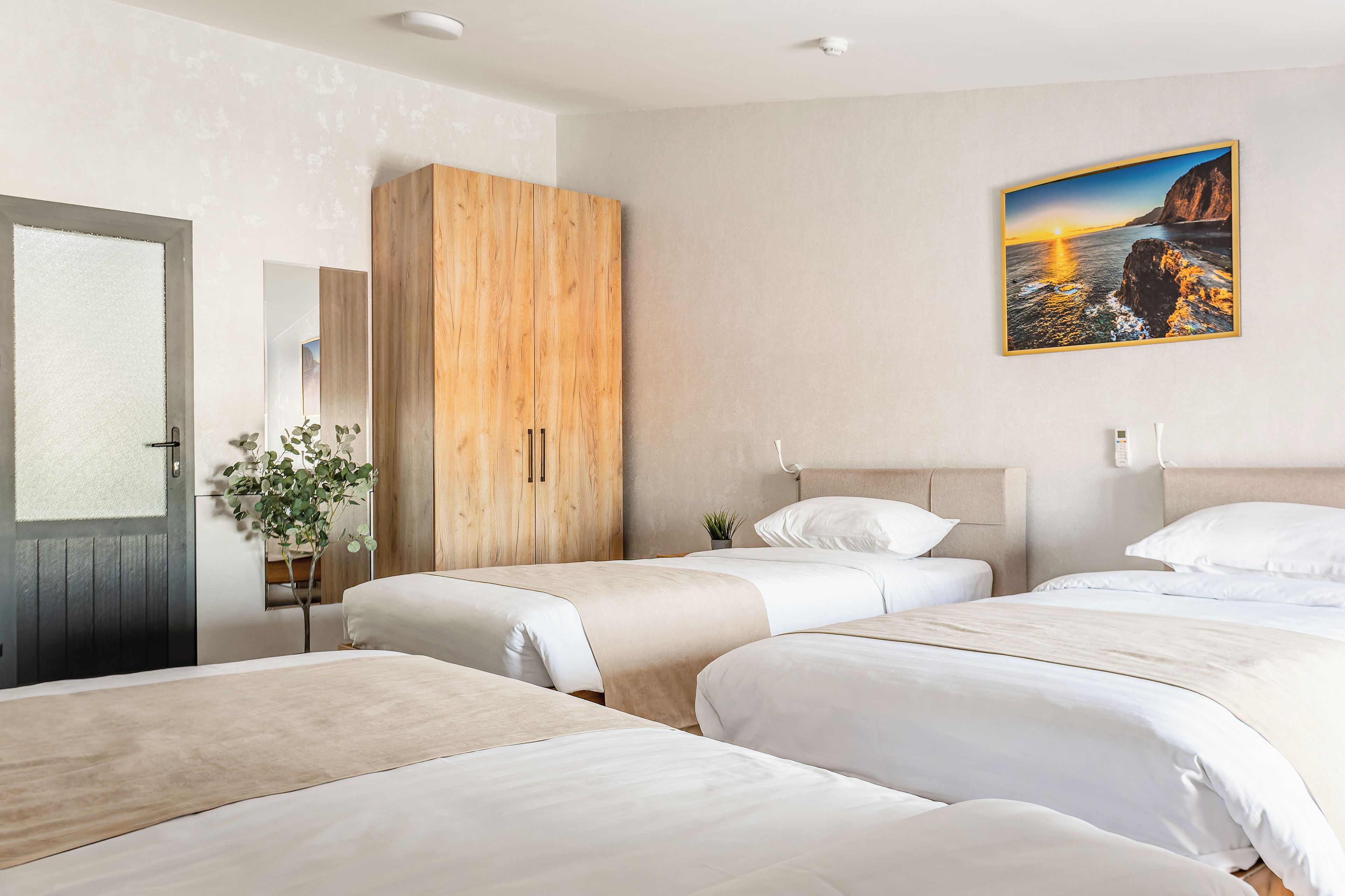 Habitación de hotel con tres camas, un armario de madera, una puerta con vidrio esmerilado y un cuadro con una escena costera.