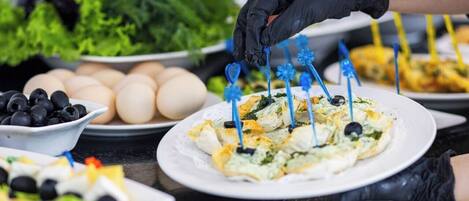 Uma pessoa servindo comida, com um prato de aperitivos, incluindo itens com palitos azuis e uma tigela de azeitonas pretas.