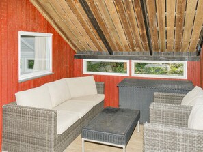 A covered outdoor seating area with wicker furniture, a red wall, and two windows with views of greenery.