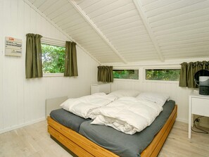 Ein Schlafzimmer mit einem hölzernen Bett, zwei fensterbepflanzten Fenstern, einer weißen Decke und einem an der Wand befestigten Thermostat.